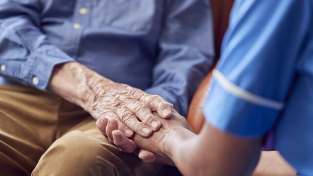 Close Up Of Female Care Worker In Uniform Holding Hands Of Senior Man Sitting In Care Home Lounge