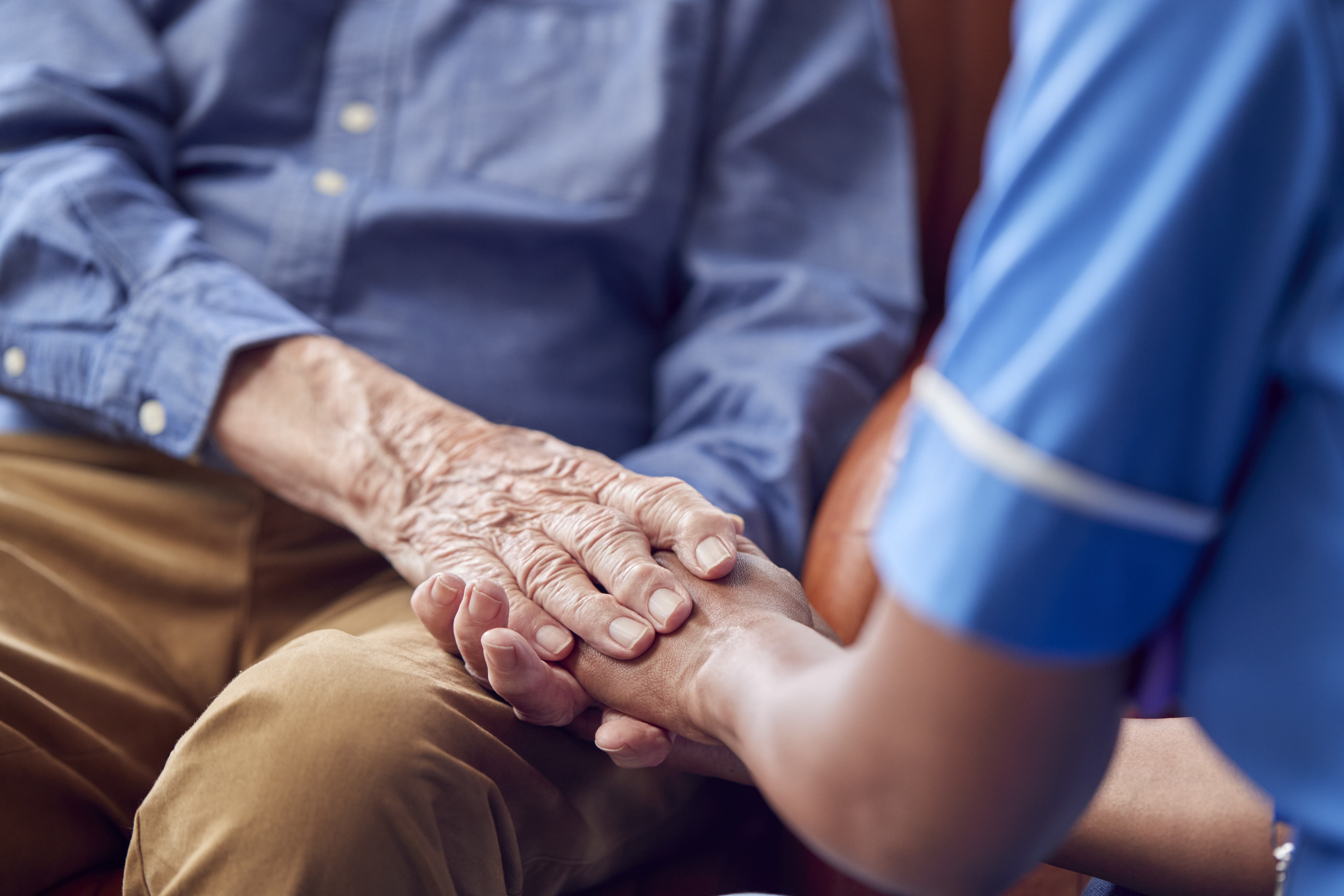 Close Up Of Female Care Worker In Uniform Holding Hands Of Senior Man Sitting In Care Home Lounge