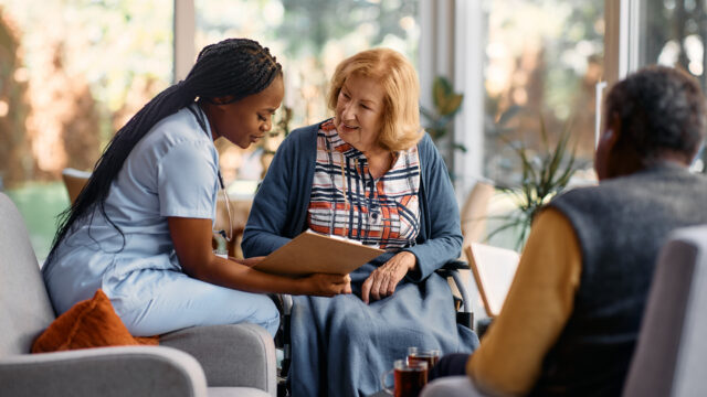 Happy Senior Woman Going Through Her Medical Data With Young Nurse At Residential Care Home.